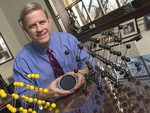 Kevin Hemker, seated between models representing how atoms are packed within an individual grain in a material, holds a silicon wafer onto which nanocrystalline aluminum thin film specimens have been deposited. Photo: Will Kirk/Homewoodphoto.jhu.edu