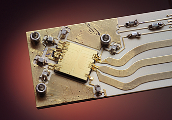 Gold ion trap on aluminum nitride backing. In NIST microwave quantum computing experiments, two ions hover above the middle of the square gold trap, which measures 7.4 millimeters on a side. Scientists manipulate and entangle the ions using microwaves fed into wires on the trap from the three thick electrodes at the lower right.
Credit: Y. Colombe/NIST