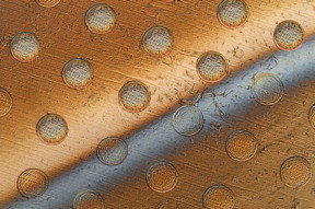 This photo shows circles on a graphene sheet where the sheet is draped over an array of round posts, creating stresses that will cause these discs to separate from the sheet. The gray bar across the sheet is liquid being used to lift the discs from the surface.

Image: Felice Frankel